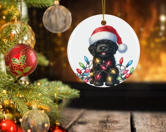 Puede incluir: Adorno navideño redondo con un cachorro negro con un gorro de Papá Noel y luces navideñas de colores. El adorno cuelga de un hilo dorado, con un árbol de Navidad y otros adornos en el fondo.