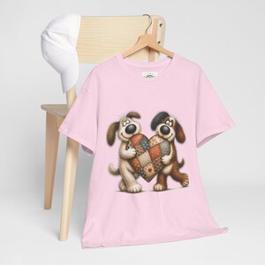 Peut inclure: Un t-shirt rose clair à manches courtes orné d'une illustration graphique représentant deux chiens de dessin animé debout côte à côte, tenant un grand cœur en patchwork multicolore. Le t-shirt est présenté sur un cintre en bois.