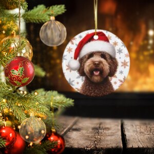 May include: A round ceramic Christmas ornament featuring a portrait of a brown curly-haired dog wearing a red and white Santa hat. The background of the ornament is white and decorated with a pattern of small grey and brown snowflakes.