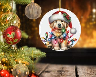 May include: A round Christmas ornament featuring a golden retriever puppy wearing a Santa hat and wrapped in colorful Christmas lights. The ornament hangs from a gold string, with a Christmas tree and other ornaments in the background.