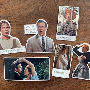 May include: A collection of stickers featuring scenes and quotes from the movie "The Sound of Music." The stickers include images of characters and phrases like "Oh help" and "Spiders??!!" on a wooden surface.