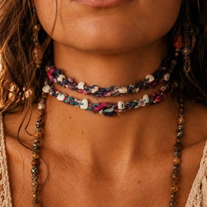 May include: A multi-strand choker necklace featuring braided fabric in shades of magenta, teal, and dark blue, accented with small, irregular white stone beads. Two long, dangling beaded necklaces with brown and metallic spacers hang below.