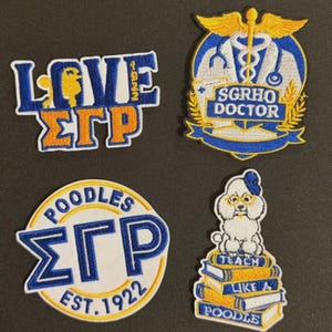 May include: Four embroidered patches with blue, orange, and white details. One patch reads "LOVE ΣΓΡ 1922" with a poodle graphic. Another features a medical symbol and the text "SGRHO DOCTOR". A third says "POODLES ΣΓΡ EST. 1922". The last shows a poodle on books.