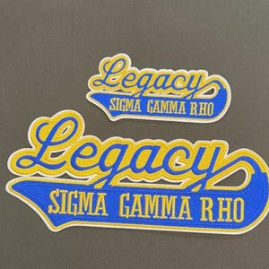 May include: Two embroidered patches with the word "Legacy" in yellow script over a blue banner with "SIGMA GAMMA RHO" in yellow. The patches have a white border and are on a gray background.