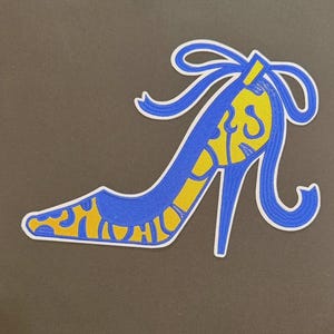 May include: A blue and yellow high-heeled shoe patch with a bow detail. The shoe features a stiletto heel and a decorative design in yellow. The patch is outlined in white and set against a dark background.