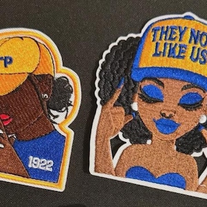 Op de afbeelding: Twee geborduurde patches met zwarte vrouwen. Eén draagt een gele pet met "ΣΓΡ" en "1922". De andere patch toont een vrouw met blauwe lippen en de tekst "THEY NOT LIKE US" op haar pet. Beide hebben een witte rand.
