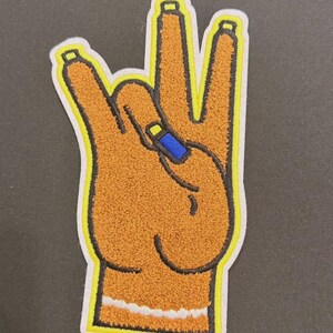 May include: Embroidered patch featuring a hand gesture with the index and pinky fingers extended. The patch has a textured, orange-brown appearance with a yellow border and a white outline. A blue nail is visible on the middle finger.