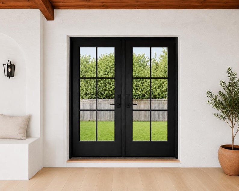 May include: Black double doors with a grid pattern, offering a view of a green lawn and trees. The doors are set in a white wall with a wooden beam above. A small potted plant sits to the right, and a wall sconce is on the left.