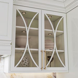May include: White kitchen cabinet with glass doors and decorative white trim. The cabinet features interior lighting and gold-toned handles. Inside, there are shelves displaying decorative items in neutral tones.