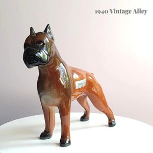 May include: A brown ceramic Boxer dog ornament with a glossy finish. The dog has a black face and ears, with a label that reads "Baron Jr.". The figurine is on a white surface. The text "1940 Vintage Alley" is in the background.