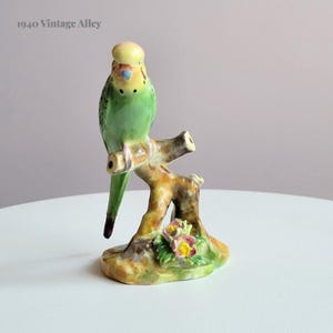May include: A green and yellow ceramic figurine of a parakeet perched on a branch with pink flowers. The figurine is on a white surface. 1940 Vintage Alley.