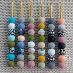 Könnte beinhalten: Eine Sammlung von Häkelnadeln mit goldfarbenen Metallhaken und farbenfrohen Perlengriffen. Die Griffe weisen eine Vielzahl von Farben und Mustern auf, darunter Türkis, Grau, Rosa und florale Designs. Die Haken sind vertikal vor einem neutralen Hintergrund angeordnet.