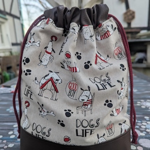 Könnte beinhalten: Ein beiger Kordelzugbeutel mit braunem Boden und Oberteil. Die Tasche ist mit Cartoon-Hunden, Pfotenabdrücken und den Worten "DOGS LIFE" und "GOOD BOY" gemustert. Der Kordelzug ist dunkelrot.