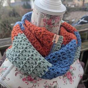 Könnte beinhalten: Ein gehäkelter Infinity-Schal in Blautönen, Grün und Orange. Der Schal wird auf einer Puppe mit Blumenmuster präsentiert. Der Schal hat ein kleines Leder-Etikett mit dem Wort "doop" darauf.