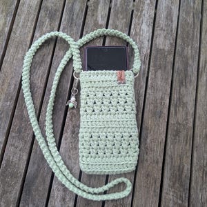 Könnte beinhalten: Eine hellgrüne gehäkelte Handytasche mit einem langen Riemen. Die Tasche hat ein kleines Leder-Etikett mit dem Wort "Fuzzy" darauf. Das Handy ist in der Tasche sichtbar.