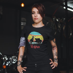 May include: A black t-shirt with a vintage-style graphic of a Vespa scooter in front of a sunset. The text "Vespa" is printed below the scooter.