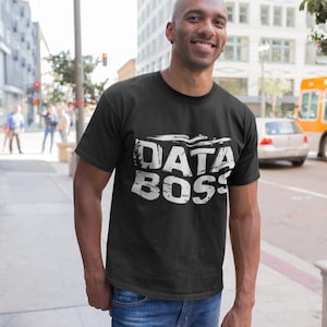 Può includere: Una maglietta nera con una grafica bianca che dice "DATA BOSS" in un font usurato.