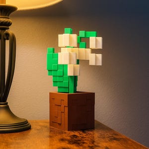 May include: A pixelated flower sculpture in a brown square pot. The flower has green stems and leaves with white flower blooms. The sculpture is on a wooden table next to a lamp with a beige lampshade.