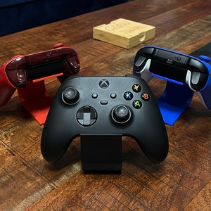 May include: Three video game controllers, one black, one red, and one blue, are displayed on a wooden surface. Each controller is in a stand.