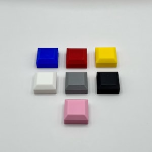 May include: Seven small, square objects in various colours: blue, red, yellow, white, grey, black, and pink. Each has a slightly angled top edge. The objects are arranged in a circular pattern against a plain background. The colours are vibrant and distinct.