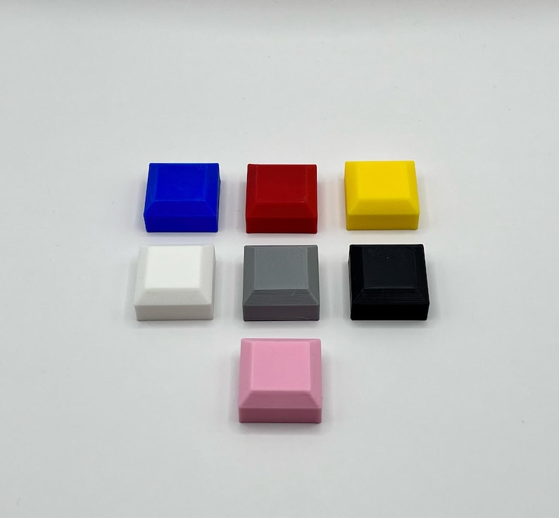 May include: Seven small, square objects in various colours: blue, red, yellow, white, grey, black, and pink. Each has a slightly angled top edge. The objects are arranged in a circular pattern against a plain background. The colours are vibrant and distinct.