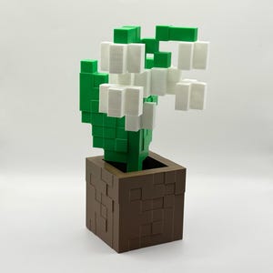 May include: A 3D-printed, pixelated flower arrangement. The flowers are white, the leaves are green, and the pot is brown. The design is reminiscent of a video game aesthetic, with a blocky, geometric style.