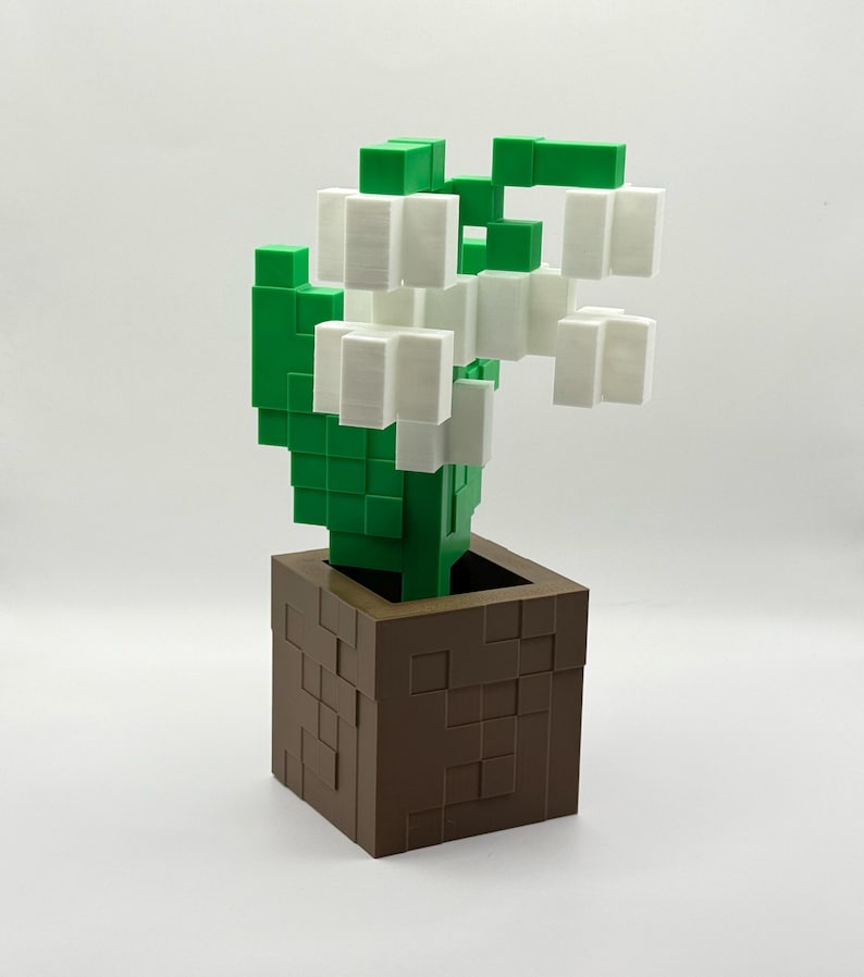 May include: A 3D-printed, pixelated flower arrangement. The flowers are white, the leaves are green, and the pot is brown. The design is reminiscent of a video game aesthetic, with a blocky, geometric style.