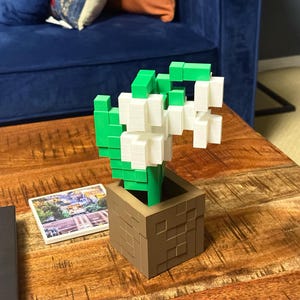 May include: A 3D-printed flower arrangement in a brown square pot. The flowers are white and the leaves are green. The pot is on a wooden table. A small picture is next to the pot.
