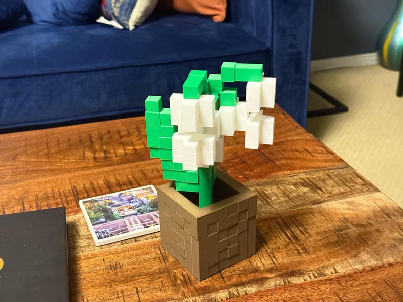 May include: A 3D-printed flower arrangement in a brown square pot. The flowers are white and the leaves are green. The pot is on a wooden table. A small picture is next to the pot.