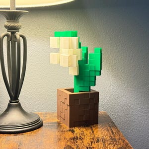 May include: A 3D-printed cactus in a brown square pot. The cactus is made of green and cream-coloured blocks, giving it a pixelated appearance. The pot has a textured surface. The item is on a wooden table.