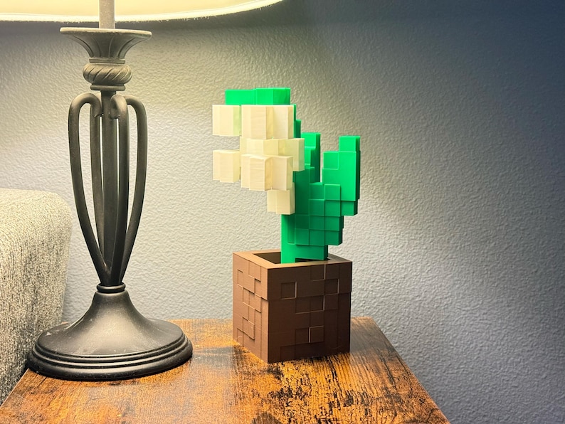 May include: A 3D-printed cactus in a brown square pot. The cactus is made of green and cream-coloured blocks, giving it a pixelated appearance. The pot has a textured surface. The item is on a wooden table.