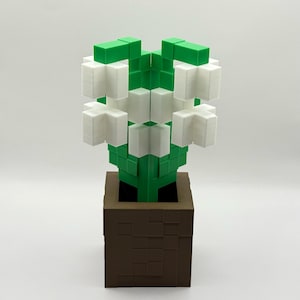 May include: A 3D-printed flower sculpture in a brown square pot. The flower has white petals and green leaves and stem. The pot has a textured, blocky design. The sculpture is set against a white background.