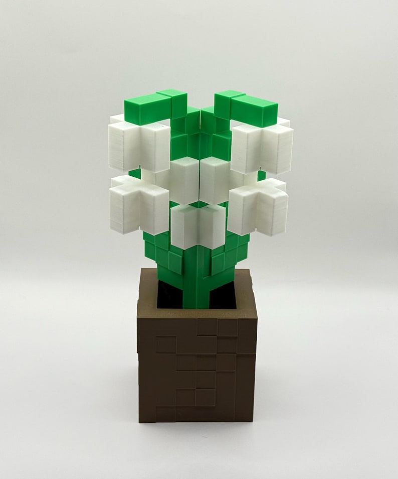 May include: A 3D-printed flower sculpture in a brown square pot. The flower has white petals and green leaves and stem. The pot has a textured, blocky design. The sculpture is set against a white background.