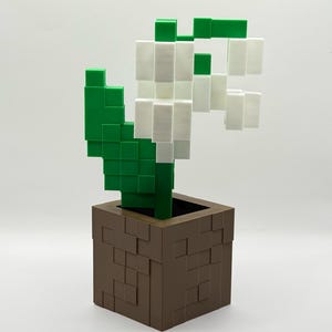 May include: A 3D-printed, pixelated lily of the valley in a brown square pot. The flower has white blossoms and green leaves and stem. The pot has a textured, blocky design. The overall aesthetic is reminiscent of a video game.