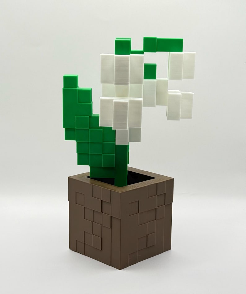 May include: A 3D-printed, pixelated lily of the valley in a brown square pot. The flower has white blossoms and green leaves and stem. The pot has a textured, blocky design. The overall aesthetic is reminiscent of a video game.