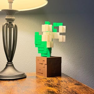 May include: A pixelated cactus sculpture in a brown pot. The cactus is made of green and white blocks, and the pot has a textured appearance. The sculpture is on a wooden surface, with a lamp and a couch in the background.