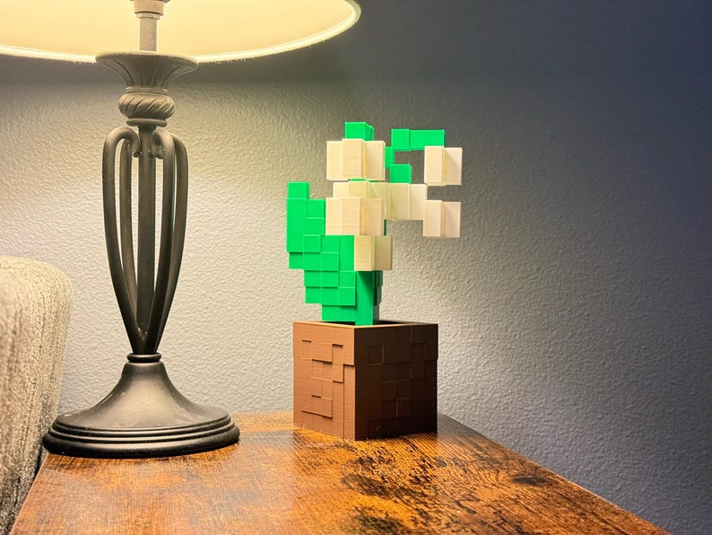 May include: A pixelated cactus sculpture in a brown pot. The cactus is made of green and white blocks, and the pot has a textured appearance. The sculpture is on a wooden surface, with a lamp and a couch in the background.