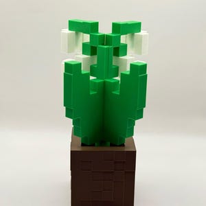 May include: A green and white pixelated cactus sculpture on a brown square base. The cactus has a geometric design, with the green sections forming the body and the white sections at the top. The base is a dark brown colour.