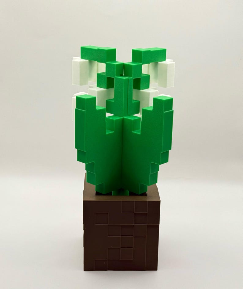 May include: A green and white pixelated cactus sculpture on a brown square base. The cactus has a geometric design, with the green sections forming the body and the white sections at the top. The base is a dark brown colour.
