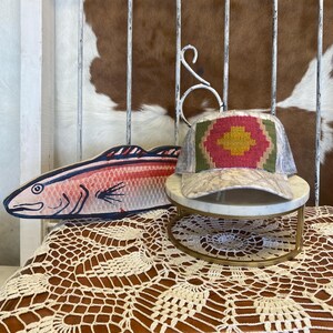 May include: A baseball cap featuring a woven geometric panel in red, yellow, and green on the front, paired with a light-colored marbled pattern on the brim and sides. Beside it sits a flat, wooden fish cutout painted in shades of pink and blue.