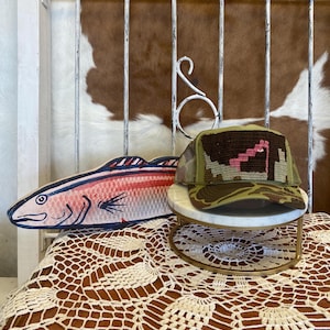 May include: A baseball cap with a camouflage brim and a brown woven front panel featuring a pink staircase design, positioned next to a flat, wooden, fish-shaped cutout painted in shades of pink, red, and blue with black outline details.