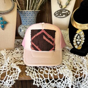 May include: A pink trucker hat with a white front panel and a brown and pink geometric design. The hat is displayed on a wooden shelf with other jewelry and decorative items.