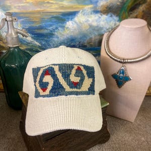 May include: Cream-colored corduroy baseball cap with a woven design in blue and red. The cap is displayed on a wooden surface, with a vintage soda siphon and a necklace with a turquoise pendant in the background.