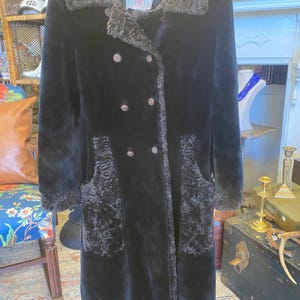 May include: A vintage black faux fur coat with a double-breasted design. The coat features a textured collar and pocket accents, with large buttons. The coat is hanging on a wooden hanger.