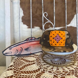 May include: A camouflage trucker hat featuring a square orange and pink crocheted granny square patch on the front panel, paired with a flat-style wooden cutout of a fish painted in shades of pink, blue, and black with visible line details.