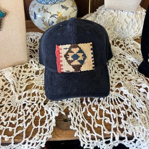 May include: Navy blue corduroy baseball cap with a rectangular patch featuring a geometric design in red, beige, and blue. The cap is displayed on a wooden surface with a white lace doily.