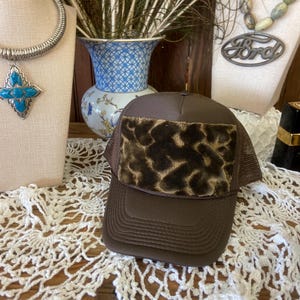 Puede incluir: Gorra de camionero marrón con un parche con estampado de leopardo. La gorra tiene una parte trasera de malla y una visera curva. La gorra se exhibe sobre un tapete de encaje blanco.
