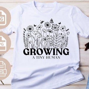 Op de afbeelding: Wit T-shirt met een zwarte lijntekening van bloemen en de tekst "GROWING A TINY HUMAN".