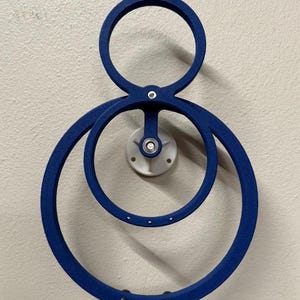May include: A blue metal kinetic sculpture with three concentric circles. The sculpture is mounted on a white wall with a silver screw.