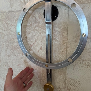 Op de afbeelding: Een zilverkleurig, cirkelvormig metalen object met een verticale, rechthoekige steun en een messingkleurig gewicht aan de onderkant. Het cirkelvormige deel heeft gelijkmatig verdeelde gaten en messingkleurige accenten. Het is gemonteerd op een zwarte cirkelvormige basis.